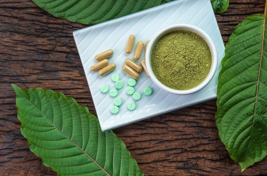 Mitragyna speciosa or kratom leaves with medicinal products in pills, capsules and powder in white ceramic bowl and wooden table, top view
